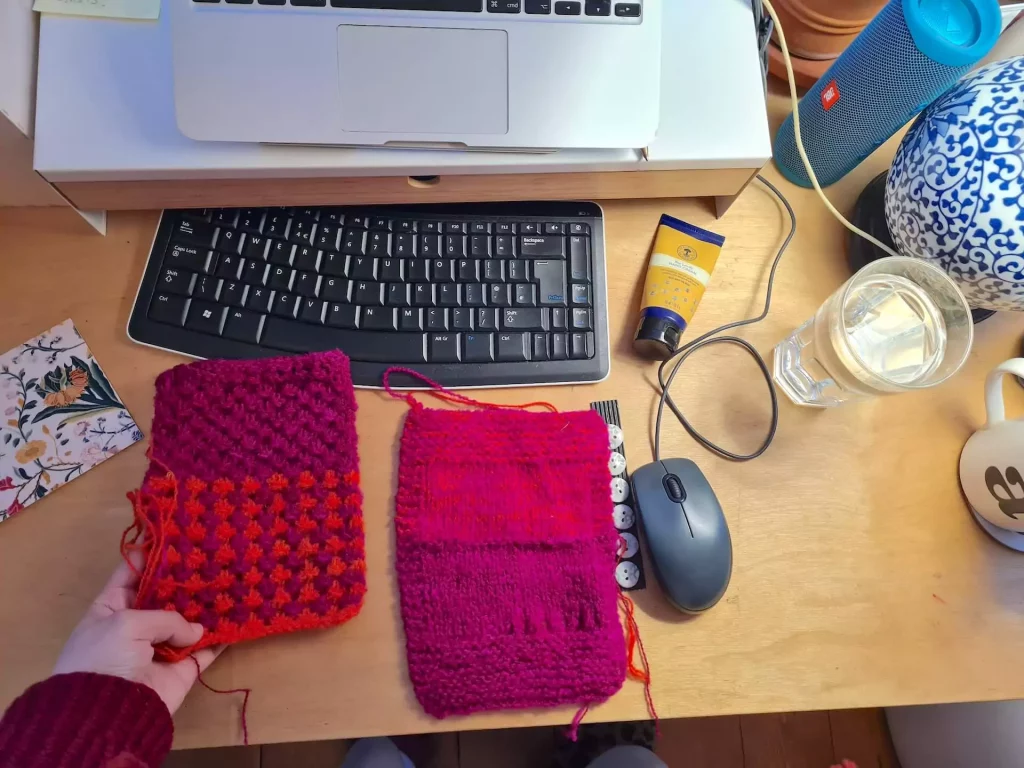 View of a desk with a hand touching swatches of knitting, a computer and coffee