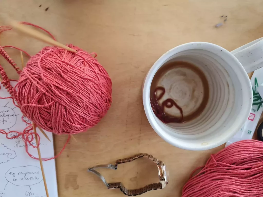 Red yarn and knitting next to a sketchbook and an empty coffeecup with yarn inside