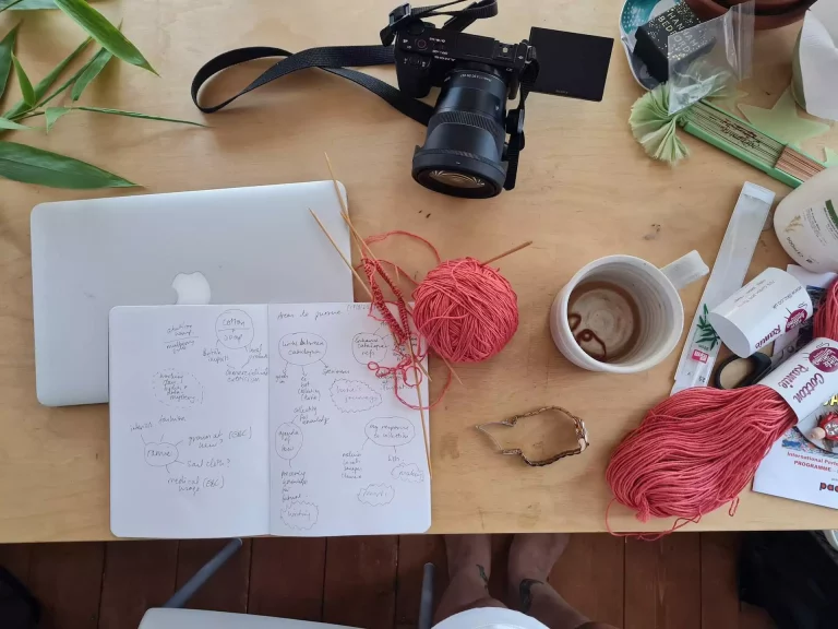 A messy creative workspace with sketchbook, laptop, knitting, red yarn, a camera and coffee